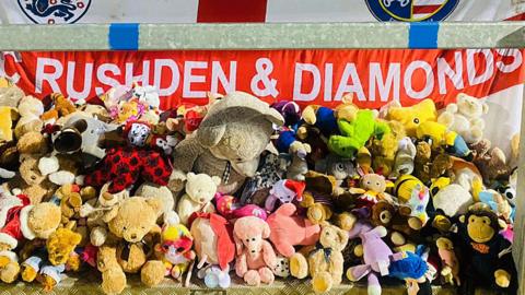 A mound of teddy bears and other soft toys, backed by an England flag with Rushden & Diamonds in white capitals across the horizontal red stripe.