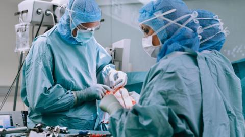 A group of doctors wearing blue scrubs, masks and hairnets. They are in a surgical theatre performing a knee replacement operation on a patient.