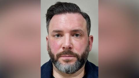A police mugshot of a man with black hair and a black beard with some grey hairs. He is facing the camera and is wearing a dark blue top.