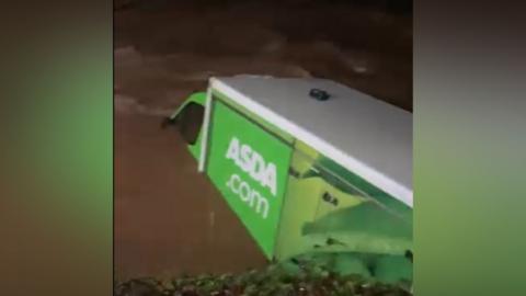 A green food delivery van that says ASDA.com on the side is trapped in brown floodwater which has rise up to the bottom of the passenger side window