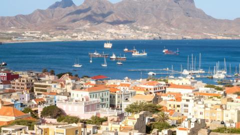 View over Mindelo, Sao Vicente, Cape Verde showing buildings with terracotta-coloured roofs in front of the blue ocean, which has several boats in it. Behind the ocean is a grey mountain and the sky is light and blue.