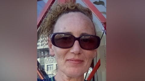 Close up of Susan Proudfoot's face. She is wearing large black sunglasses and has her hair up. She is smiling and behind her is a window with a reflection of a sign that says "Apo". She is wearing a dark striped top.