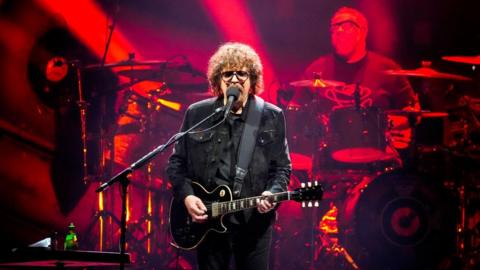 A man with long curly brown hair in dark clothing, holding a black guitar in front of a band lit with red lights