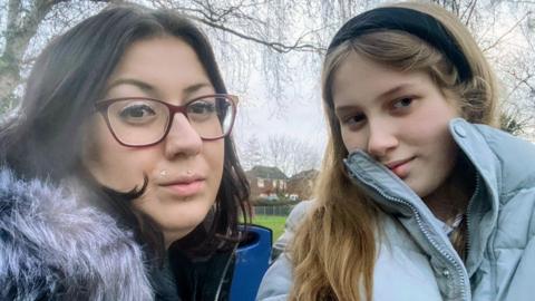 Selfie-style picture of Rhiannon Van Der Zanden and her daughter Alexis. Rhiannon has black hair and glasses and wears a coat with a fur collar. Alexis has long blonde hair worn with a black alice band. Her coat is light blue. 