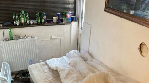 A bedroom in the illegally converted bungalow, showing an unmade bed, an electrical heater, a radiator and peeling faded cream wallpaper.