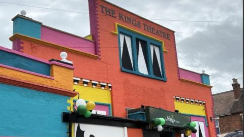 A building painted orange, blue. pink, yellow and with black and white accents. The words "The Kings Theatre" are printed at the top. Balloons are attached to the door. 