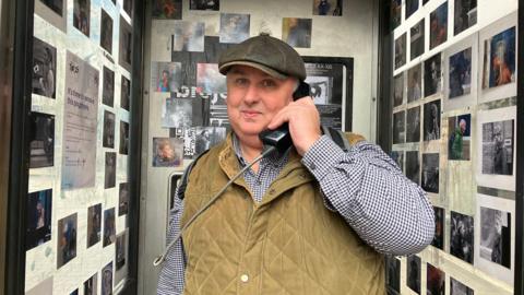 A man in a stone gilet, checked shirt and hat smiles as he holds a phone box phone to his ear. Behind him and to each sides are photos stuck onto the walls of the phone box.