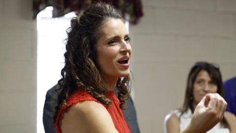 Julia Letlow is seen talking to constituents which smiling and appearing to talk. She is turned to the side and is seen from the shoulder up. She is wearing a red top.