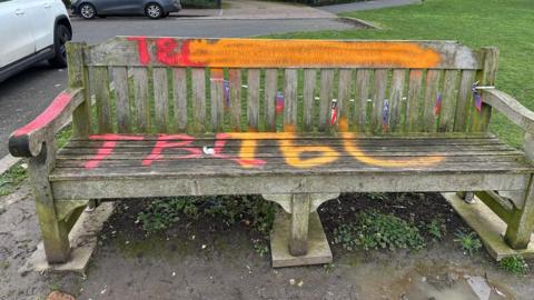 A wooden bench is standing on concrete next to a small patch of grass. It has been spray painted with graffiti.