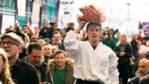 Smithfield meat trader holds a large piece of meat on his head as he he is surrounded by many customers