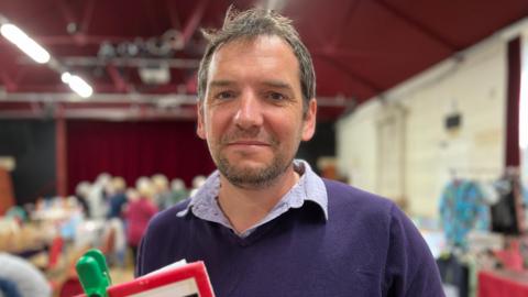 Man wearing blue jumper and blue shirt holding a clipboard