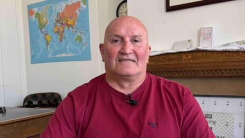 Councillor Watkins looks at the camera. He is wearing a red T-shirt and has a shaved head.