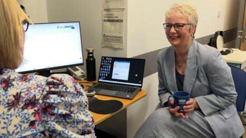 Karin Smyth is sitting in a GP consultation room holding a cup of tea. She is speaking to a GP who is sitting slightly out of frame.