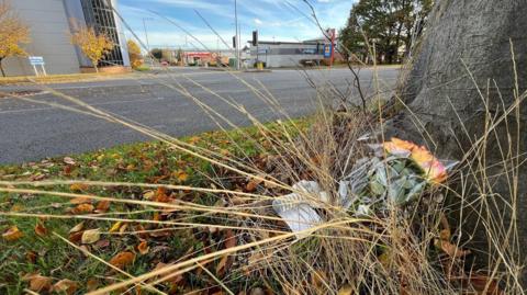 A bunch of flowers lie next to a tree, nearly buried under the dying stalks of grass. The tree is next to a road and beyond that are industrial buildings.