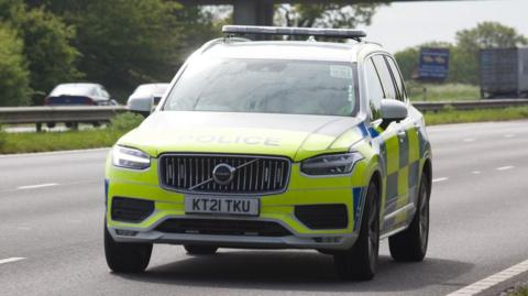 A police car is parked on a three-carriageway road. It is yellow and blue. The rest of the lanes appear to be empty 