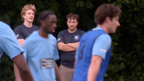 Man watches footballer on sideline.