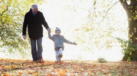 An elderly gentleman takes a walk in the autumn leaves with his young grandson, while the sun shines on green leaves still clinging to the trees in the background - they are both wearing warm coats