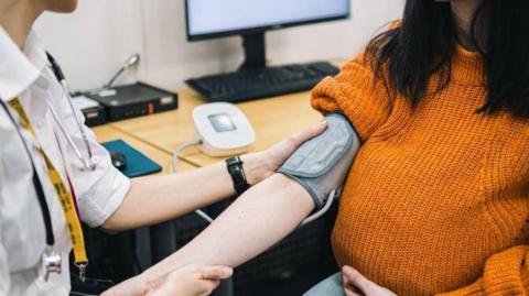 A woman extending her arm out with a doctor measuring her heart rate with a device strapped to her elbow. She is wearing an orange jumper and has long hair down to her shoulders. A desk with a computer can be seen in the background.