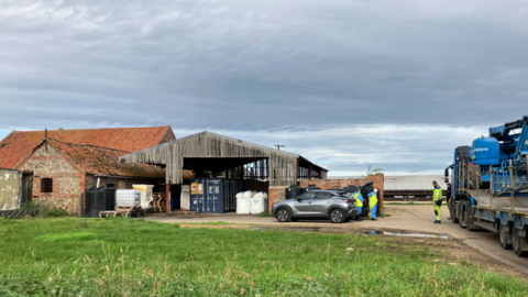 Three people wearing hi-visibility uniforms are shown near a car parked next to a flint and brick farm building and a wooden barn, with a large HGV parked nearby