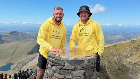 Joel Redhead with multi-colour hair and a black beard stands next to his friend Shane who is wearing a blue hat on the summit of Snowdon. The pair are wearing yellow hooded tops with the Chasing Rainbows charity logo on them.