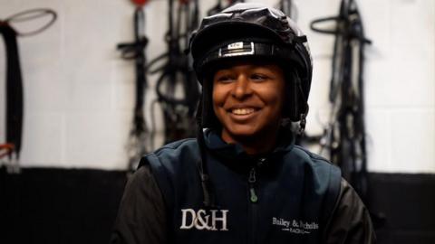 Aamilah Aswat smiles as she is pictured in her riding kit in the tack room at Kim Bailey and Mat Nicholls' training yard