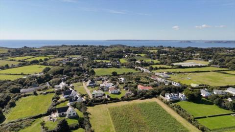 An aerial view of sark showing green fields and communities of houses. The sea, Guernsey and Herm are visible in the background. 