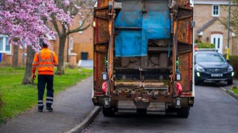 The rear of a bin lorry as it travels along a residential street. A man in a high-vis jacket walks alongside the vehicle. A blossoming tree is shown in the foreground.