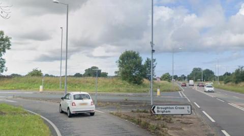 A turnoff towards Brigham from the A66. A white Fiat 500 is turning off the road. Six cars are driving in both directions along the A66 on the right.