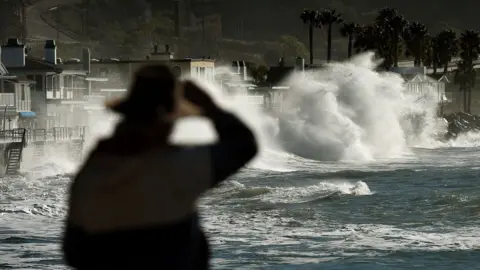 A silhouetted man holds onto his hat while looking out over waves in California during an El Niño event in 2016