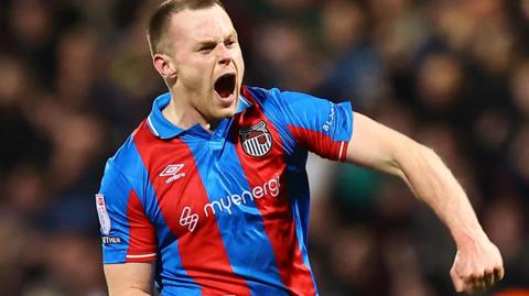 Cameron McJannet wearing a red and blue striped Grimsby away jersey celebrates with fans