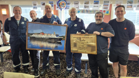 Sianar fhireannach air bòrd bàta-aiseig. Tha iad air an èideadh ann an aodach gorm a' chriutha aig CalMac. Tha iad a' cumail dealbh de bhàta-aiseig agus leac umha air a' comharrachadh gu bheil fear aca a' leigeil dheth a dhreuchd
