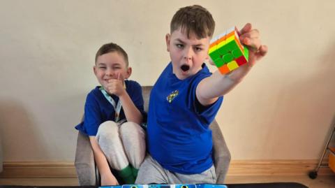 Two young boys wearing matching blue t-shirts. The younger boy is sitting in an armchair and giving a thumbs-up to the camera. The older boy is standing in front of the chair and holding up a Rubik's Cube in an heroic gesture. On a table in front of them are a selection of other toys, including several more Rubik's Cubes.