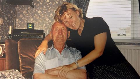 Cathy Bowles is sitting with her arm around her father with their heads close together. They are both smiling   