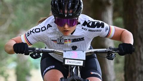 A woman on a mountain bike with helmet and glasses and a white top is tucked low over the handlebars with mud spattered on her.