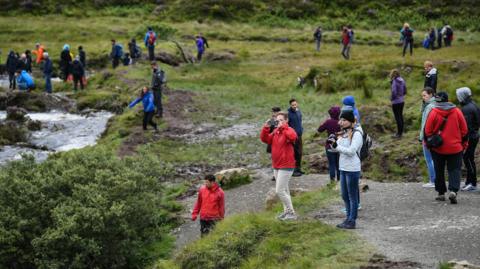 A picture taken on Skye in 2017 showing tourists wearing brightly coloured jackets and fleeces exploring a glen with a river running through it.