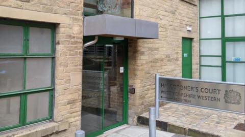 The outside of a coroner's court, as indicated by a silver sign at the front of the building. The edges of the building's glass doors are green.