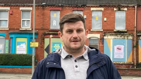 Tom Murphy, who has brown hair brushed to one side and is wearing a blue coat over a grey polo-neck t-shirt, stands in front of terraced houses which have their windows boarded with metal sheets