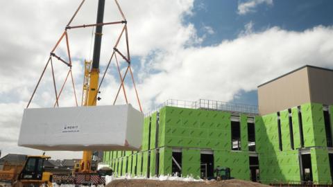 A building site featuring a large crane-like structure lowering a huge block to the ground next to a metal frame construction covered in green cladding