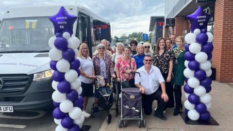 A large group of happy looking people stand beside a white mini bus. There are purple and white balloons making two columns either side of the group with a star-shaped balloon at the top of each column reading "SERVICE ROUTE 66". A retail park is behind them with shops seen.