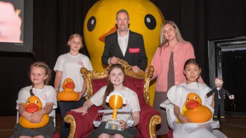 Four girls are sitting on chairs, wearing white t-shirts and holding yellow rubber ducks. They are smiling next to Alex Horne who is wearing a white shirt and a black blazer. He is standing in front of a giant yellow rubber duck. To the right of the girls is a woman with long blonde hair wearing a pink jacket and a dress.