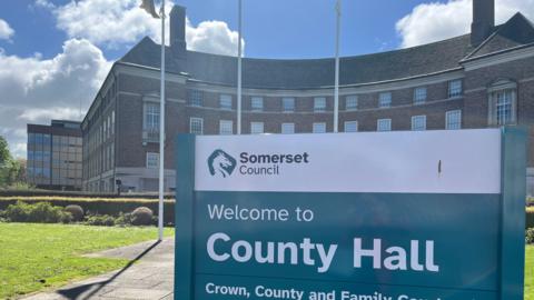 A sign in front of Somerset Council County Hall, which is a red brick building in the background.