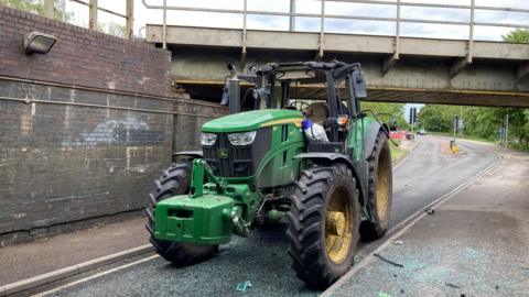 A green John Deere tractor has a badly damaged roof and wing mirrors. It is on a road under a bridge and there is debris and shattered glass on the road and pavement.