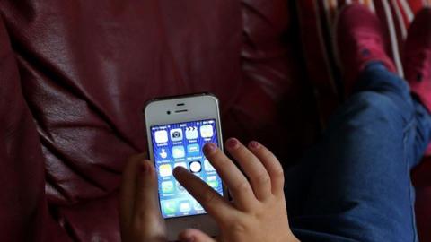 A child's hand with rough red nail varnish pointing towards an icon on a mobile phone. The child is wearing red trainers and is sitting on a brown chair.