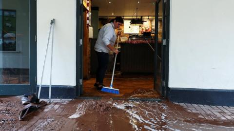 A woman sweeps water away from a coffee shop after severe flooding caused by Storm Claudia, in Monmouth, Wales