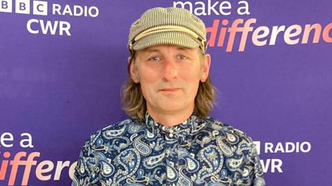 A man with a flat cap and a blue buttoned-up shirt, stands in front of a purple background which says BBC Radio CWR, Make a Difference.