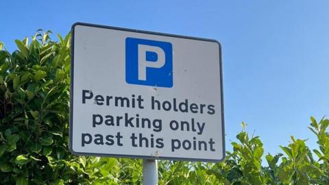 A road sign has a blue 'P' on it and reads 'Permit holders parking only past this point'. It is in front of a green, leafy hedge under a clear blue sky.