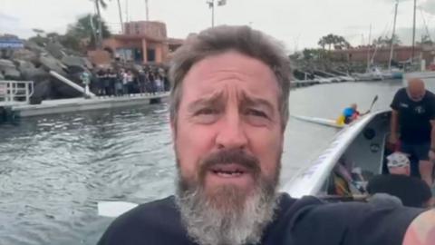 Andy Merry taking a selfie off the side of a rowing boat with other rowers behind him in a harbour.  He has a thick greying beard.