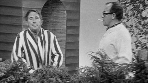Black and white still from a TV broadcast, showing Ernie Wise in a dressing down and Eric Morecambe in a cardigan, both stood behind a waist high hedge