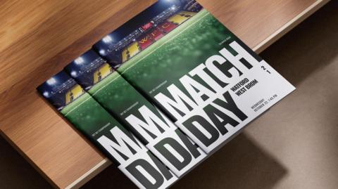 A stack of football programmes rests on a wooden surface. The covers feature a football stadium at the top with bright floodlights and yellow and red seating. Below the stadium photo, large bold text reads “MATCH DAY".