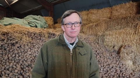 A man with glasses and brown hair combed to the left is wearing a tweed jacket and standing in front of a mound of potatoes.